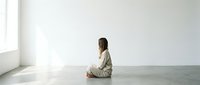 Young woman sitting cross-legged on floor in minimalist white room practicing meditation or mindfulness in peaceful morning light from window.