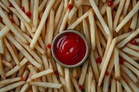 Golden crispy french fries scattered across frame with small metal bowl of bright red ketchup in center, overhead view of classic fast food combination.