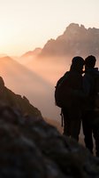 Silhouette of couple hikers embracing at mountain summit during golden hour sunrise with layered peaks and warm atmospheric haze in background.