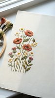 Hand embroidered wildflower bouquet with poppies, daisies and thistles on natural linen fabric with colorful threads and embroidery hoop nearby.
