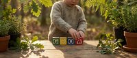 Child arranging colorful alphabet blocks spelling GROW on wooden table surrounded by potted plants in sunny garden setting for educational development.