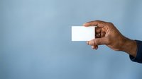 Male hand with dark skin tone holding blank white business card against blue background for corporate branding and professional networking purposes.