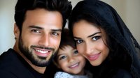 Middle Eastern family portrait with bright smiles, mother wearing traditional black hijab, father with neat beard, and cheerful young child between them against neutral background.