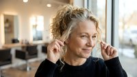Mature Caucasian woman with curly blonde hair adjusting wireless earbuds in modern office environment for technology and workplace communication.