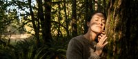 Young woman with closed eyes leaning against moss-covered tree trunk in peaceful forest setting during golden hour sunlight for wellness and mindfulness concepts.