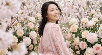 Young Asian woman in pink lace dress smiling among blooming cherry blossoms and roses. Spring portrait for lifestyle, beauty, fashion concepts.