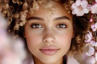 Young woman with natural curly hair and glowing skin surrounded by delicate pink cherry blossoms, expressing spring freshness and natural beauty in soft lighting.