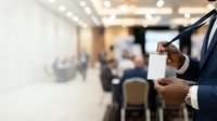Male professional in dark business suit holding blank white card at corporate conference or business event with blurred attendees in background.