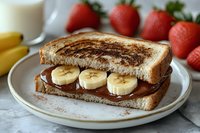 Grilled sandwich with chocolate spread and banana slices on plate with fresh strawberries and milk glass for breakfast meal preparation.