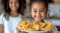 Cheerful young girl with curly hair holding plate of crispy potato chips, with another child blurred in background. Healthy snack concept.