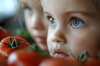 Close up portrait of young caucasian child with bright blue eyes gazing intently past fresh red tomatoes in foreground, creating artistic depth of field effect for food and lifestyle content.
