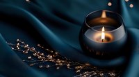 Glowing candle in glass jar on dark teal silk fabric with decorative golden branch, creating cozy atmospheric mood for relaxation or meditation.