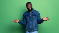 Young African American man in denim jacket shrugging with open palms against bright green background, expressing uncertainty or casual indifference.
