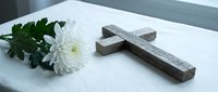 Wooden cross and white chrysanthemum flower on white surface near window creating peaceful memorial or religious meditation scene for spiritual reflection.