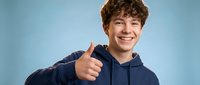 Young Caucasian teenage boy with curly brown hair wearing navy blue hoodie giving thumbs up gesture with bright smile on light blue background.
