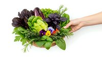 Fresh mixed salad greens and herbs in wooden bowl with woman hand reaching, healthy organic vegetables and edible flowers on white background.