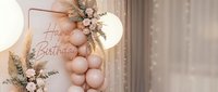 Elegant birthday party decoration with blush pink balloon arch, floral arrangement, pampas grass, and glowing lights for a boho celebration.