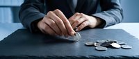 Caucasian male professional in dark business suit placing coin on table with scattered coins, representing financial planning, savings, investment strategy, and money management for corporate concepts.