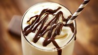 Creamy milkshake topped with swirls of dark chocolate sauce and striped straw in glass, close up view against wooden background. Indulgent dessert drink.