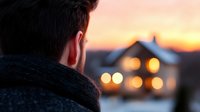 Young man in warm winter sweater looking at glowing house windows during golden sunset hour, creating cozy evening atmosphere and sense of coming home.