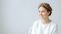 Young woman with light skin tone and blonde hair styled in a braided updo, wearing a white casual t-shirt, smiling warmly against a neutral gray background. Suitable for lifestyle, wellness, and portrait projects.