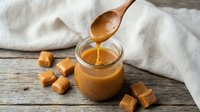 Golden caramel sauce dripping from wooden spoon into glass jar with soft caramels on rustic wood table for dessert recipes and sweet treats.