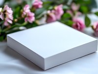 White square gift box on light surface with soft focus pink roses in background, elegant product photography setup for packaging presentation.