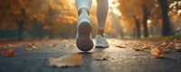 Person jogging on autumn path with fallen leaves during golden hour sunrise, athletic footwear closeup for fitness and wellness lifestyle concepts.