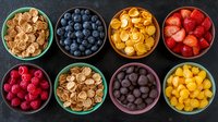 Colorful bowls filled with fresh berries, chocolate drops and crunchy mini cereal pieces arranged in rows on dark background, top view breakfast composition.