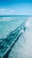 Crystal clear turquoise ocean waves gently washing onto pristine white sandy beach under bright blue sky with soft clouds for travel and vacation concepts.
