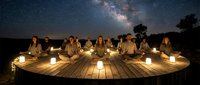 Group meditation session under starry night sky with Milky Way galaxy visible above peaceful outdoor wooden platform illuminated by warm candles.