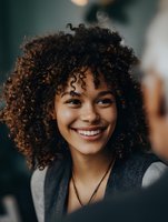 Young woman with curly hair smiling during conversation, showing genuine happiness and connection in casual social setting.