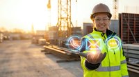 Female construction worker wearing safety helmet and reflective vest holding glowing digital sphere at sunset industrial site with cranes and building materials visible in background.