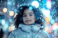 Young African American girl with curly hair wearing silver winter jacket smiles warmly against blurred bokeh lights and falling snow creating magical winter atmosphere.