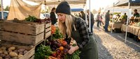 Young blonde woman in beanie and plaid shirt shopping for fresh organic vegetables at outdoor farmers market, selecting produce from wooden crates on sunny day.