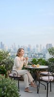 Relaxed blonde woman enjoying morning coffee on urban rooftop terrace with city skyline view, casual lifestyle moment for wellness and leisure content.