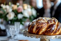 Fresh braided challah bread with golden crust on vintage silver cake stand, elegant table setting with champagne flute and soft floral arrangement in background.