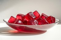 Red translucent gelatin cubes arranged in white ceramic bowl casting soft shadows on light surface, appetizing dessert with glossy texture and vibrant color.