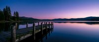 Wooden dock extending into calm lake at twilight with purple and pink sky, forested mountains in background, serene nature landscape at dusk.