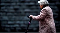 Senior woman in pink floral coat walking with mobility scooter handle against dark background, side view portrait highlighting aging and independence.