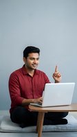 Young man with dark hair and South Asian ethnicity wearing a burgundy button-up shirt, sitting cross-legged on a cushion while using a laptop and pointing upward with one finger against a neutral gray background for business and technology concepts.
