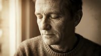 Mature Caucasian man with wrinkled face and closed eyes, wearing knit sweater, bathed in warm natural light from window, conveying contemplation, aging, and introspection for editorial and wellness content.