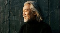 Senior Caucasian man with long gray hair and beard in black turtleneck against textured dark wall, peaceful expression with eyes closed in contemplative moment.