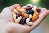 Mixed nuts and dried fruits in palm of hand against blurred green background, featuring almonds, cashews and raisins, showcasing healthy snack options.