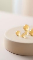 Golden gel capsules and spheres arranged on a white ceramic dish against a soft, neutral background. Suitable for healthcare, wellness, supplement, and pharmaceutical marketing materials.