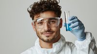 Young Hispanic male scientist in protective equipment holding test tube with blue liquid chemical solution in laboratory setting against light background.