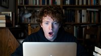 Surprised young man with curly reddish-brown hair and fair complexion working on laptop in home library setting, expressing shock or amazement at screen content.