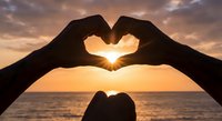Hands forming heart shape silhouette against golden sunset over ocean horizon. Romantic gesture symbolizing love, connection and unity during magical hour.