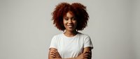 Confident young African American woman with curly hair smiling with arms crossed wearing white t-shirt against neutral background for business content.