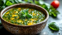 Fresh homemade pesto sauce in rustic ceramic bowl garnished with basil leaf, surrounded by green herbs and red tomato on stone surface.